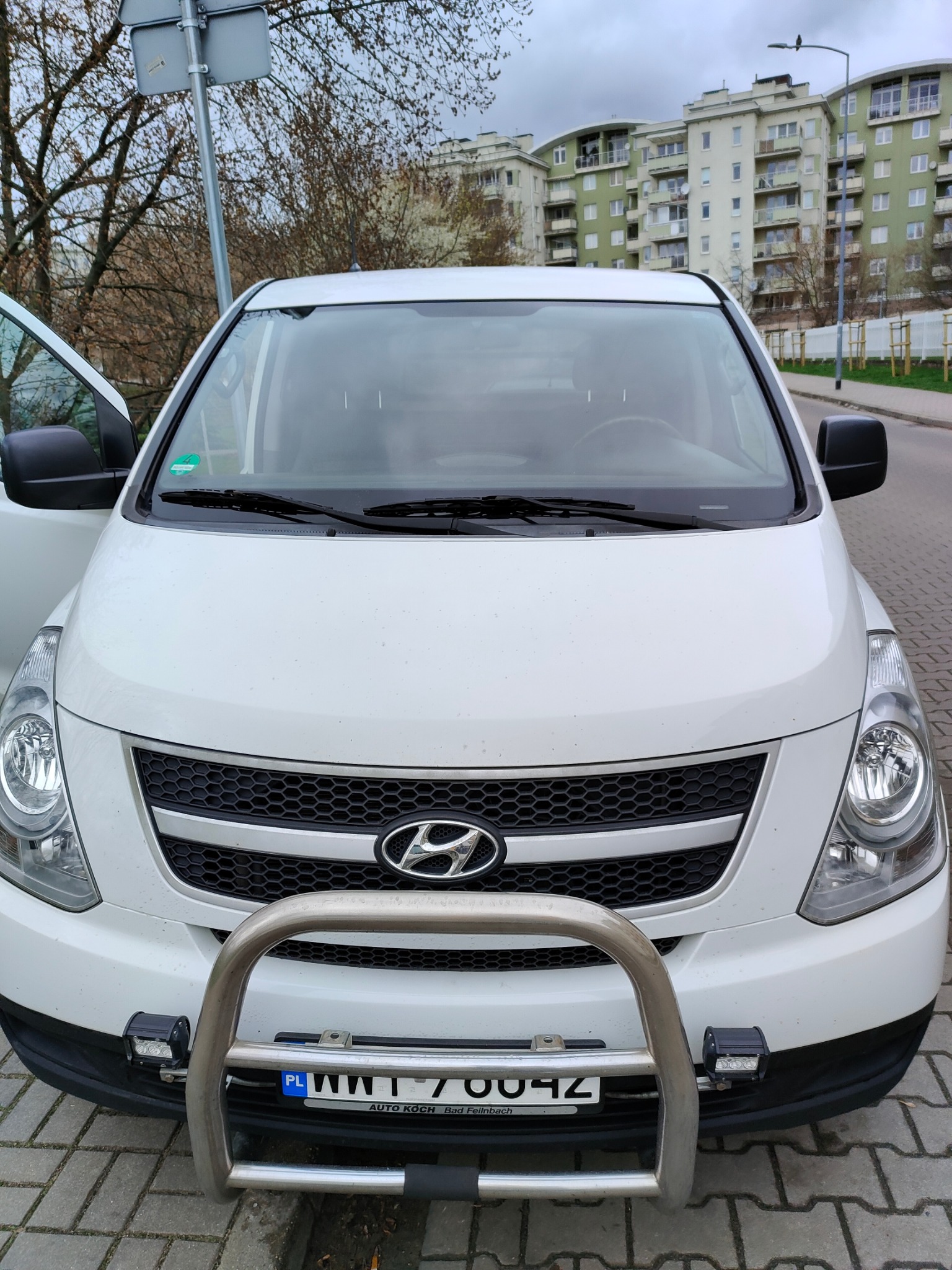 Biały Hyundai H-1 z orurowaniem i dodatkowymi reflektorami, zaparkowany na chodniku z otwartymi drzwiami kierowcy, na tle bloku mieszkalnego i drzew bez liści.