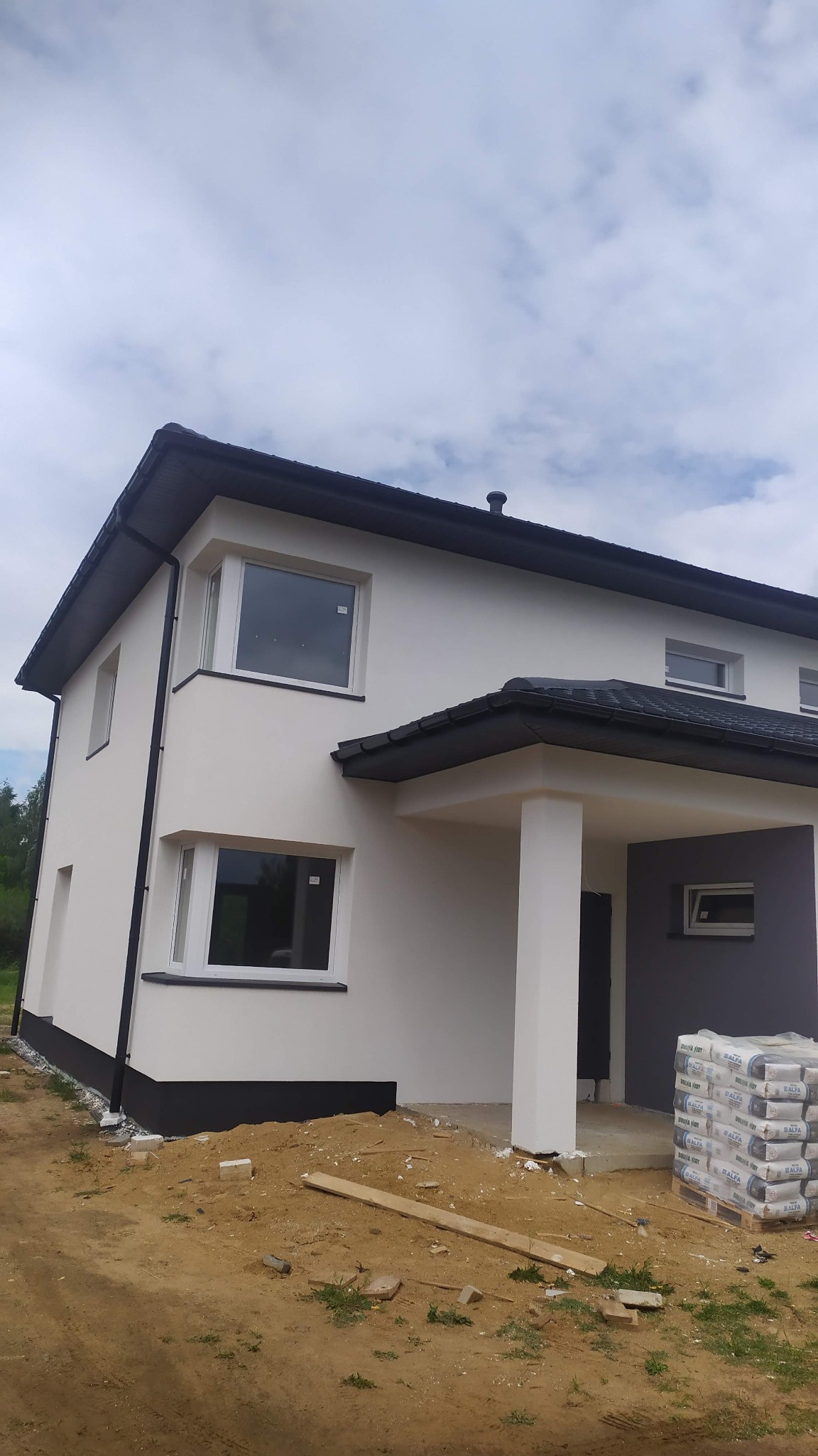 Dwukondygnacyjny dom z ocieploną elewacją w kolorze białym i szarym, widoczne worki z zaprawą na palecie, teren wokół w trakcie prac wykończeniowych.