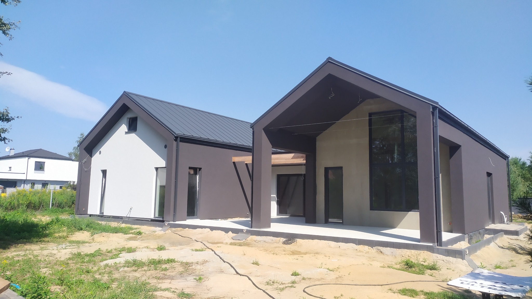 Nowoczesny dom jednorodzinny z elewacją w kolorach bieli i grafitu, widoczne elementy drewniane zadaszenia tarasu, teren wokół domu w trakcie prac wykończeniowych.