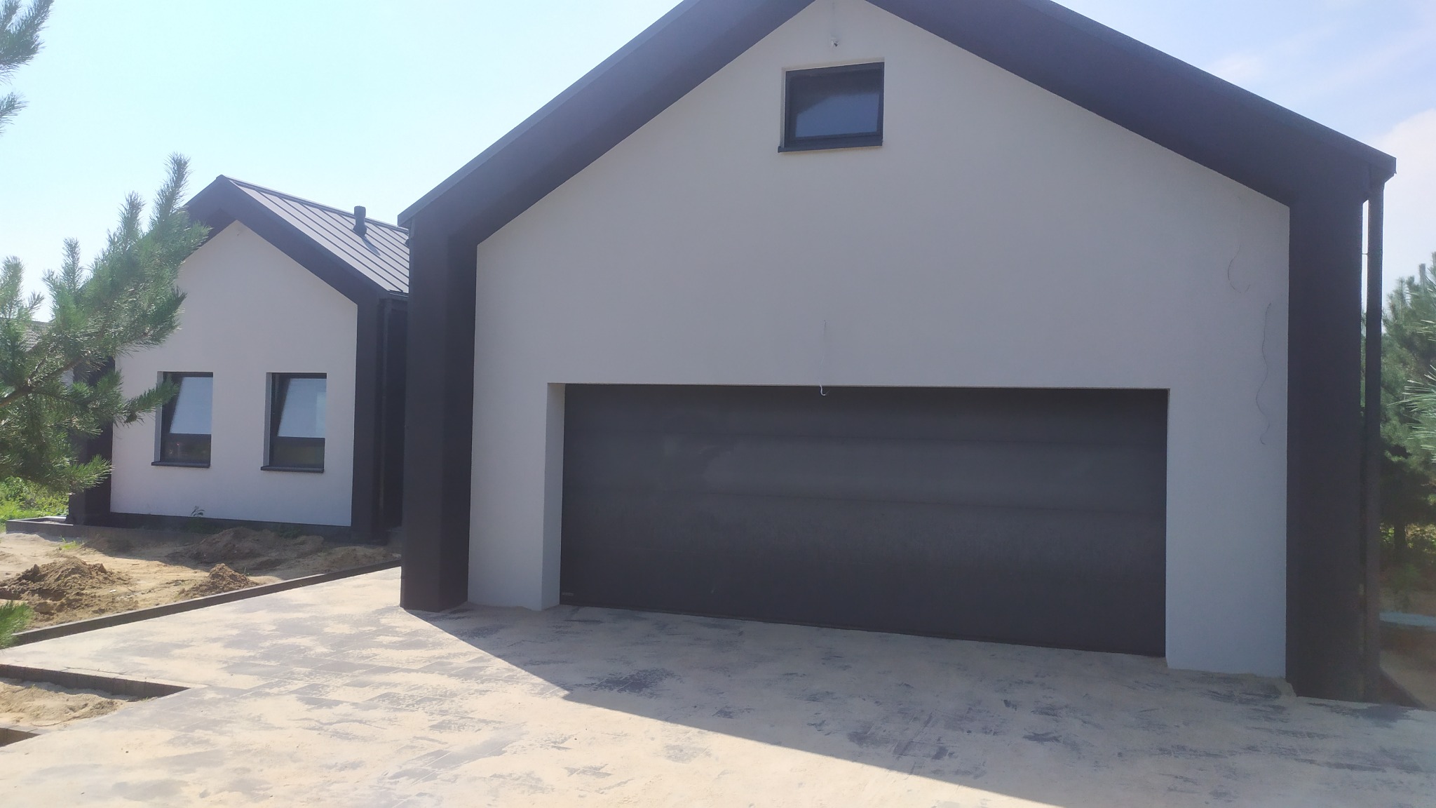 Nowoczesny dom jednorodzinny z szarą elewacją, grafitowymi elementami wykończeniowymi i bramą garażową, widok z zewnątrz w słoneczny dzień.