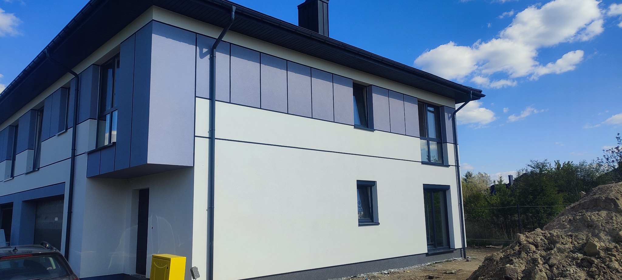 Nowoczesny budynek bliźniaczy z elewacją w kolorach bieli i szarości, wykończony panelami, widoczny fragment samochodu i żółta skrzynka na listy.