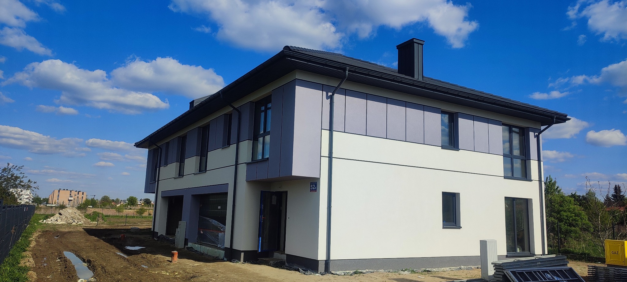 Dwukondygnacyjny dom z szarą i białą elewacją panelową, czarnym dachem i ramami okiennymi, w trakcie wykańczania terenu wokół budynku, na tle błękitnego nieba z chmurami.