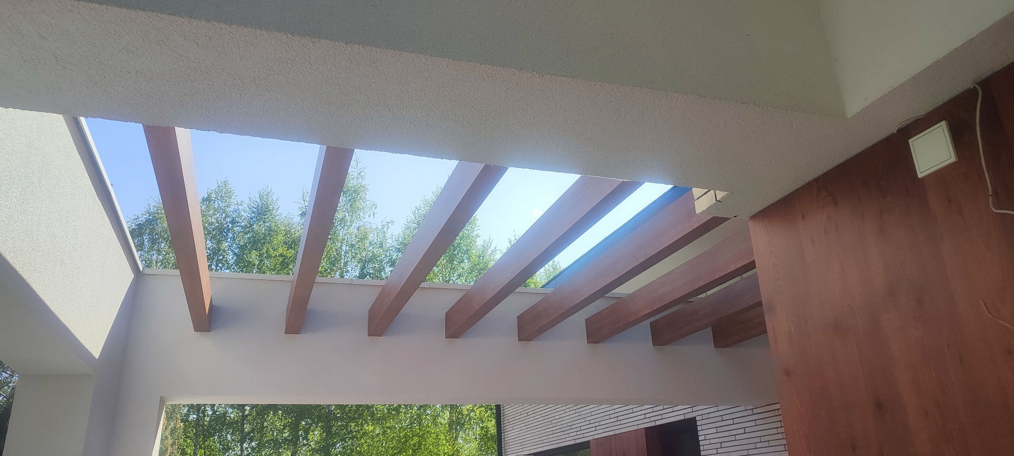 Widok z dołu na fragment elewacji z drewnianymi belkami i tynkowaną powierzchnią, błękitne niebo i zielone drzewa w tle.