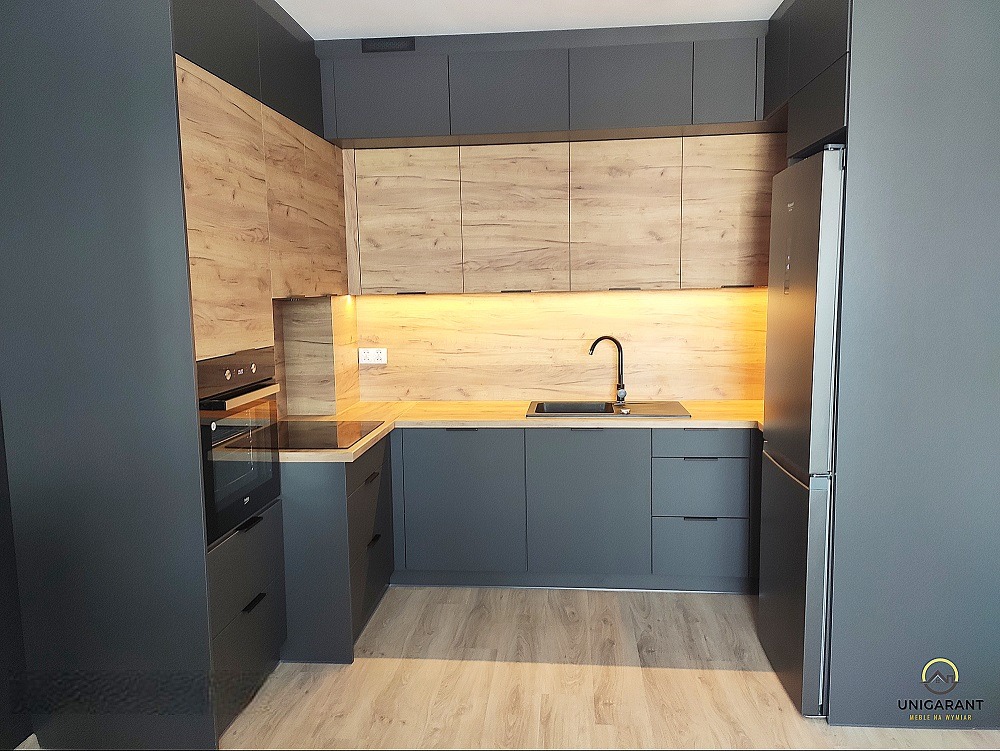 Nowoczesna kuchnia w odcieniach szarości i drewna z podświetleniem LED pod szafkami, widoczny zlew z czarną baterią i fragment lodówki.