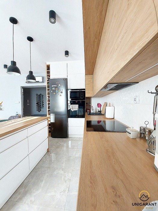 Nowoczesna kuchnia z drewnianymi blatami i białymi szafkami, oświetlona wiszącymi lampami, z widocznym fragmentem wyspy kuchennej i szarymi płytkami na podłodze.