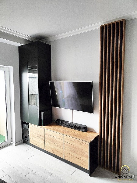 Nowoczesna zabudowa RTV w salonie: szafka z frontami imitującymi drewno, czarna szafa z lustrem i pionowy panel dekoracyjny z drewnianych listew.