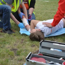Instruktorzy udzielają pierwszej pomocy osobie poszkodowanej na trawie, opatrując zakrwawioną rękę; widoczna otwarta apteczka z wyposażeniem.