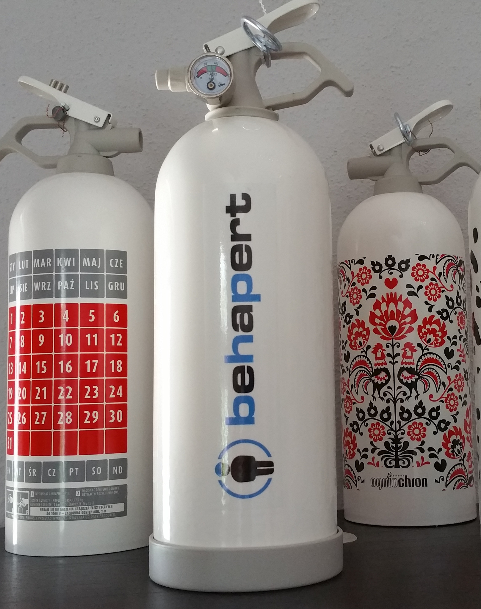 Trzy gaśnice: jedna z kalendarzem, druga z logo 'behapert', trzecia z ludowym wzorem kwiatowym i kogutami, stojące na ciemnej powierzchni.