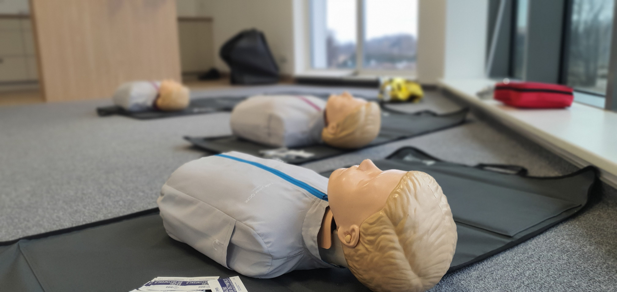 Manekiny do ćwiczeń resuscytacji na matach, w tle apteczka pierwszej pomocy i defibrylator.