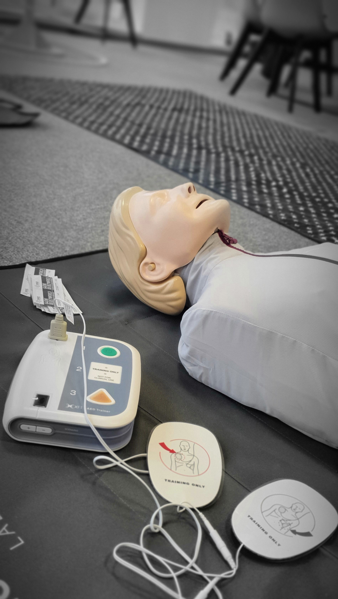 Manekin do resuscytacji z podłączonym defibrylatorem treningowym i elektrodami na czarnej macie, w tle dywan i krzesła.