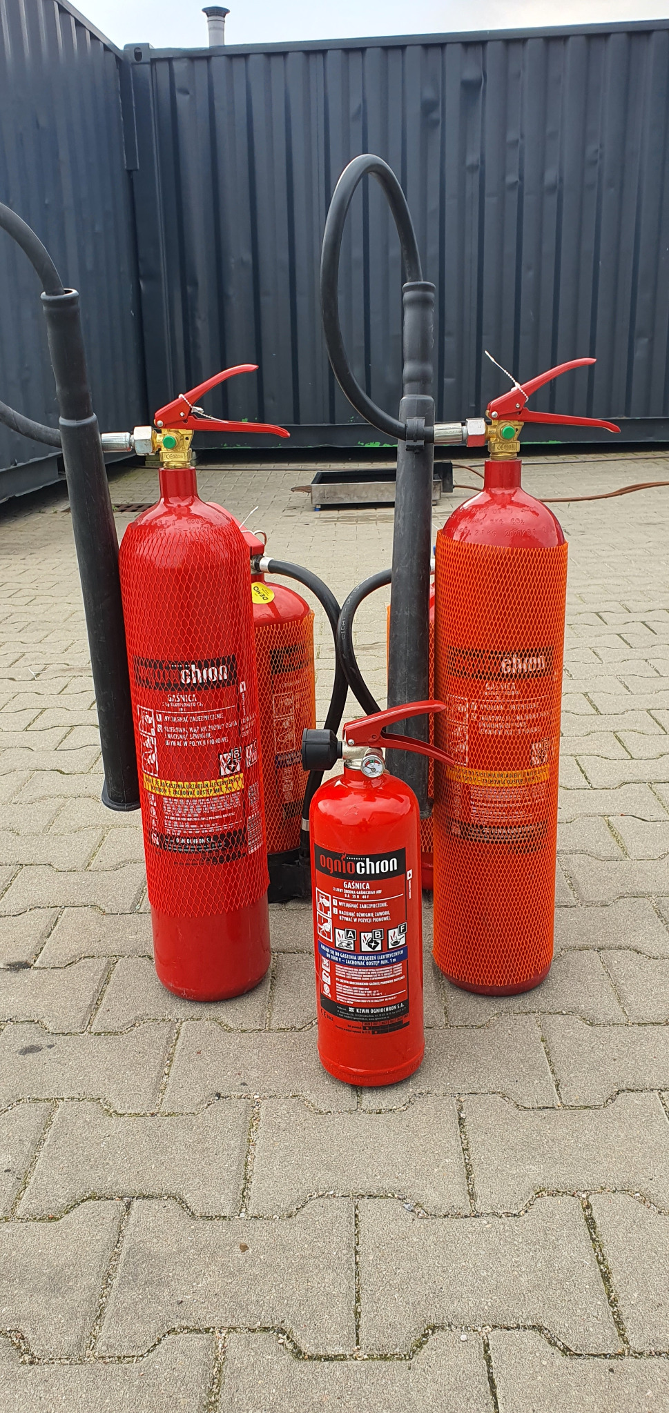 Grupa czerwonych gaśnic różnych rozmiarów, stojących na szarej kostce brukowej, na tle ciemnej, metalowej ściany kontenera.