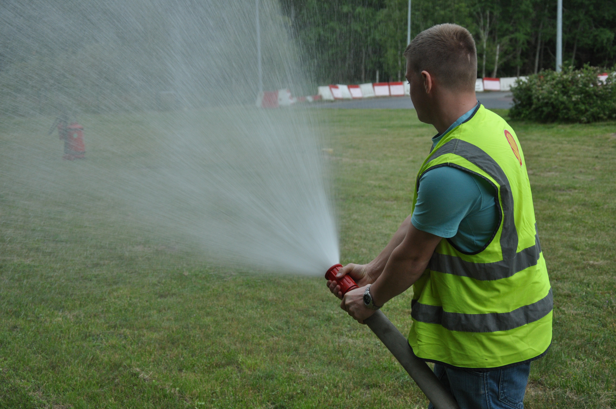 Mężczyzna w kamizelce odblaskowej obsługuje wąż strażacki, strumień wody rozpryskuje się na trawniku, w tle hydrant i drzewa.
