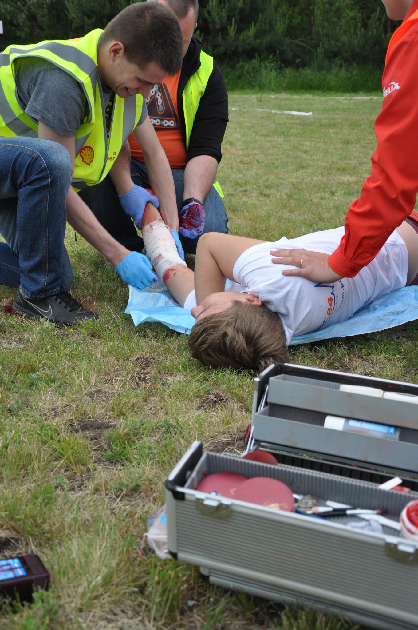 Instruktorzy udzielają pierwszej pomocy osobie poszkodowanej na trawie, opatrując zakrwawioną rękę; widoczna otwarta apteczka z wyposażeniem.