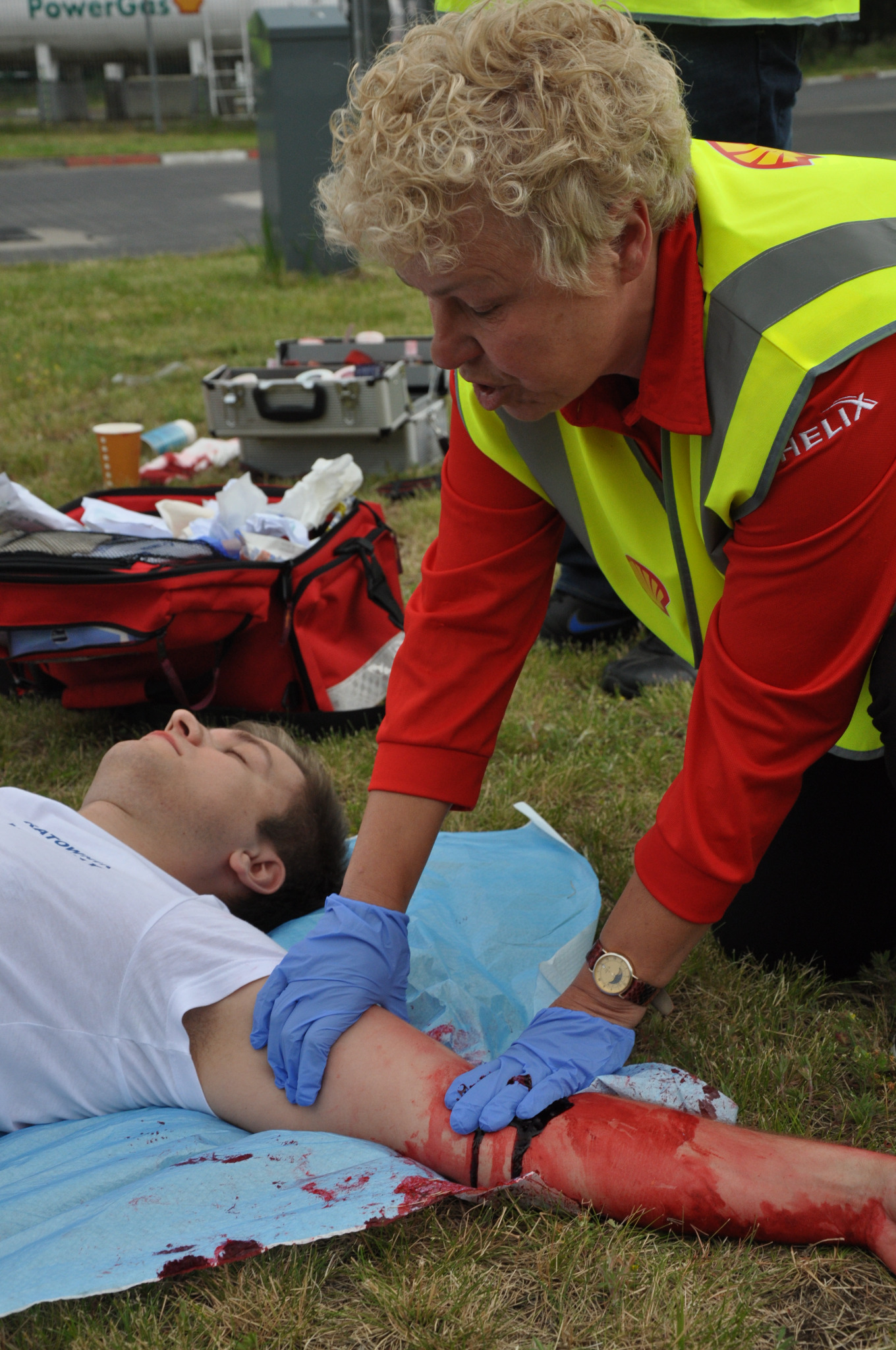 Kobieta w odblaskowej kamizelce udziela pomocy poszkodowanemu z realistyczną symulacją krwawiącej rany na ramieniu, na trawie rozłożona niebieska płachta, widoczna torba medyczna i apteczka.