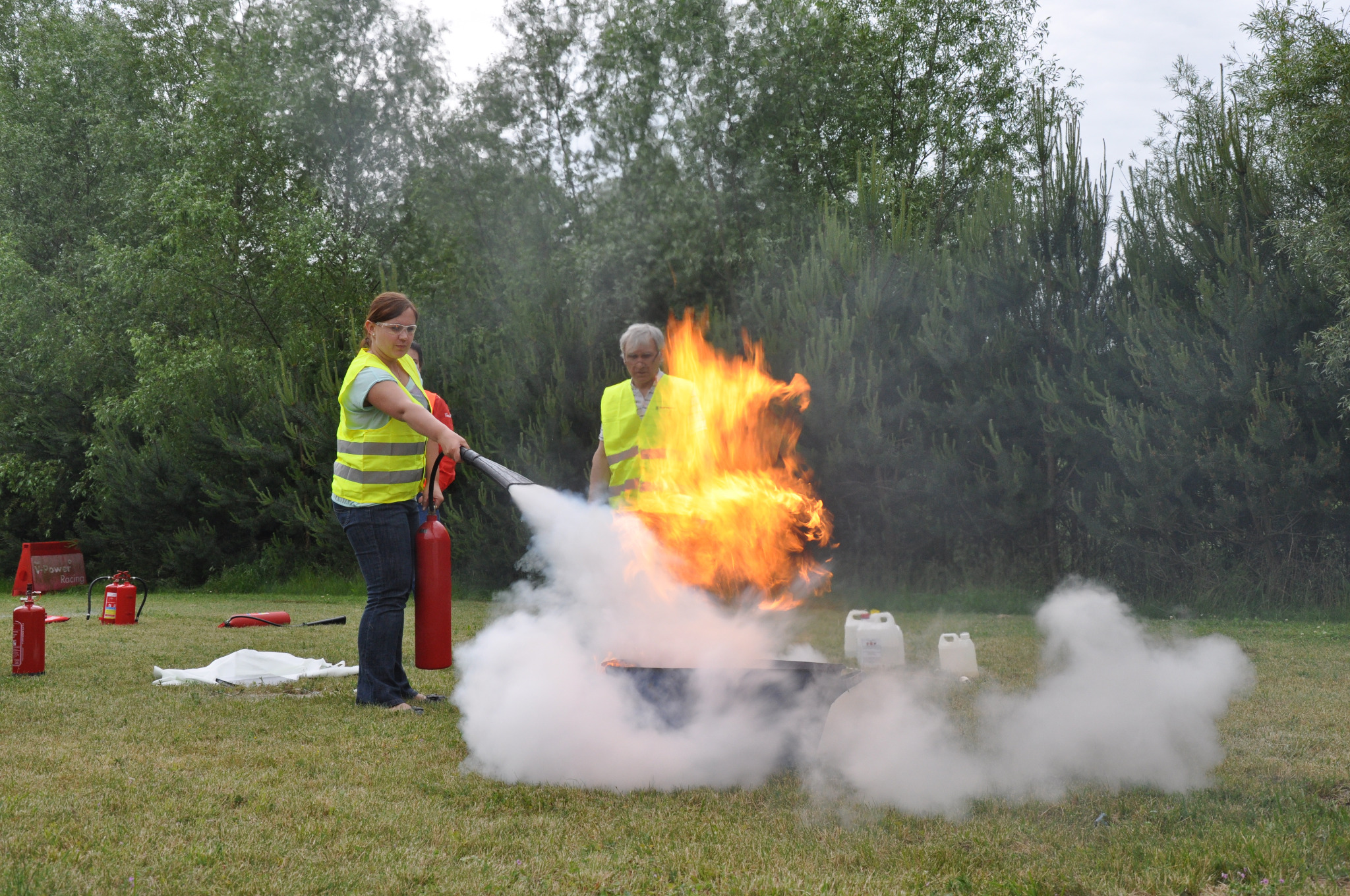 Osoba w okularach i kamizelce odblaskowej gasi płonący ogień przy użyciu gaśnicy proszkowej podczas szkolenia BHP na trawiastym terenie, z widocznymi dodatkowymi gaśnicami i manekinem w tle.