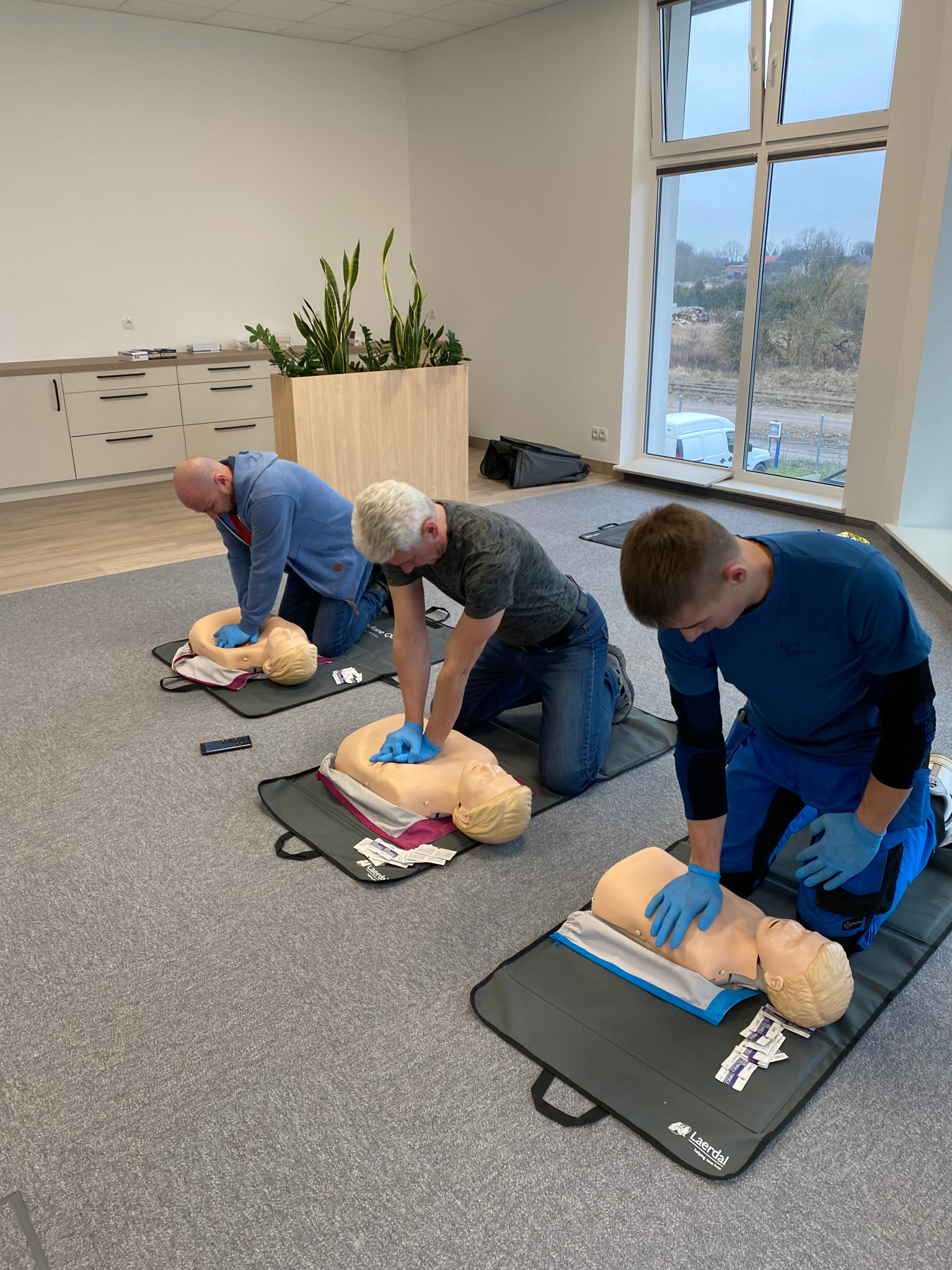 Trzech mężczyzn w niebieskich rękawiczkach klęczy obok manekinów do resuscytacji krążeniowo-oddechowej na szarym dywanie w jasnym pomieszczeniu z dużym oknem.