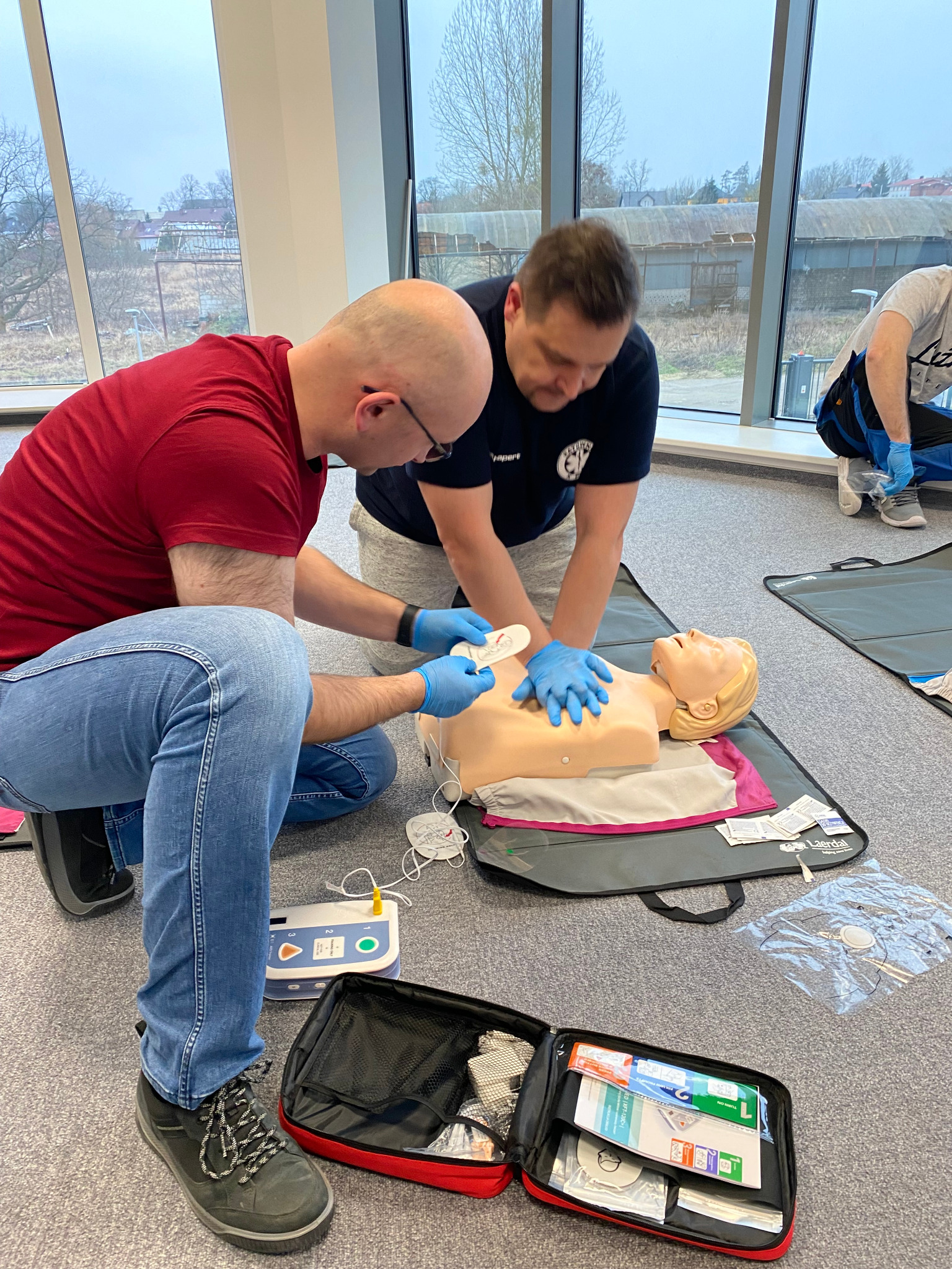 Dwaj mężczyźni w niebieskich rękawiczkach ćwiczą resuscytację krążeniowo-oddechową na manekinie, obok otwarty zestaw pierwszej pomocy i defibrylator, widok z góry, jasne pomieszczenie z dużym oknem...