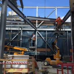 Dwóch pracowników w pomarańczowych kamizelkach na podnośnikach koszowych montuje stalową konstrukcję budynku na tle niebieskiego nieba, widoczne zabezpieczenia budowy i dźwig z łańcuchem.