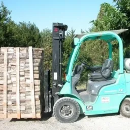 Wózek widłowy w kolorze turkusowym transportuje paletę z ułożonymi kawałkami drewna na zewnątrz, w tle zieleń drzew.