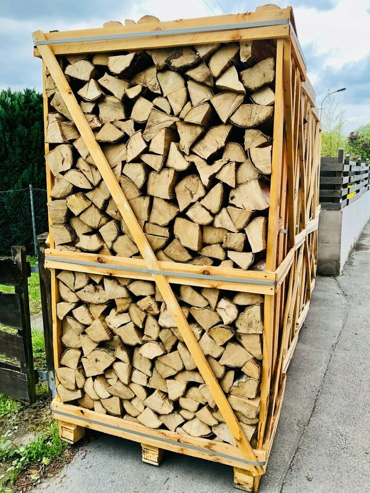 Dwa drewniane kontenery wypełnione pociętym drewnem kominkowym, zabezpieczone drewnianymi listwami, ustawione jeden na drugim na palecie, widok z boku na tle chodnika i zieleni.