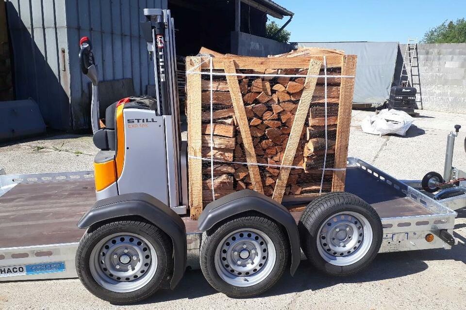 Wózek widłowy Still przewozi na przyczepie skrzynię z pociętym drewnem kominkowym, widoczny fragment budynku i drabina w tle.