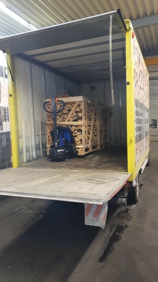 Wnętrze skrzyniowego samochodu dostawczego z otwartymi drzwiami, załadowanego paletami z pociętym drewnem kominkowym, obok wózka paletowego.