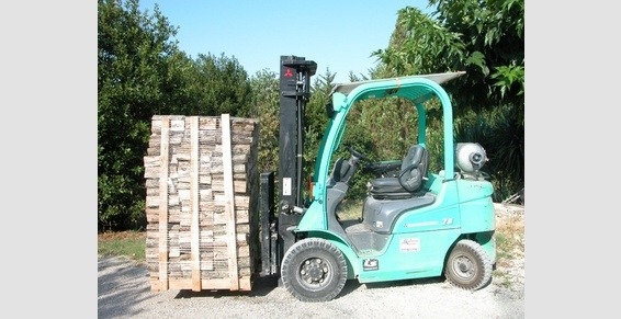 Wózek widłowy w kolorze turkusowym transportuje paletę z ułożonymi kawałkami drewna na zewnątrz, w tle zieleń drzew.