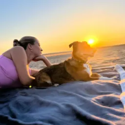 Kobieta i pies leżą na kocu na plaży, patrząc na zachód słońca.