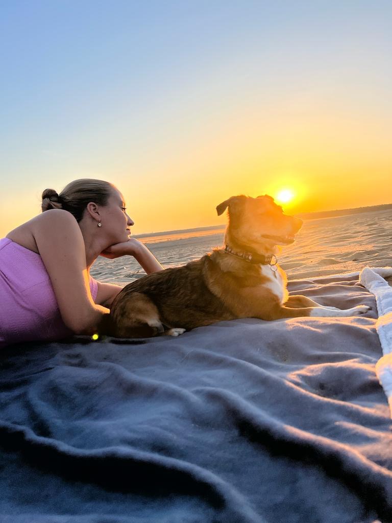 Kobieta i pies leżą na kocu na plaży, patrząc na zachód słońca.