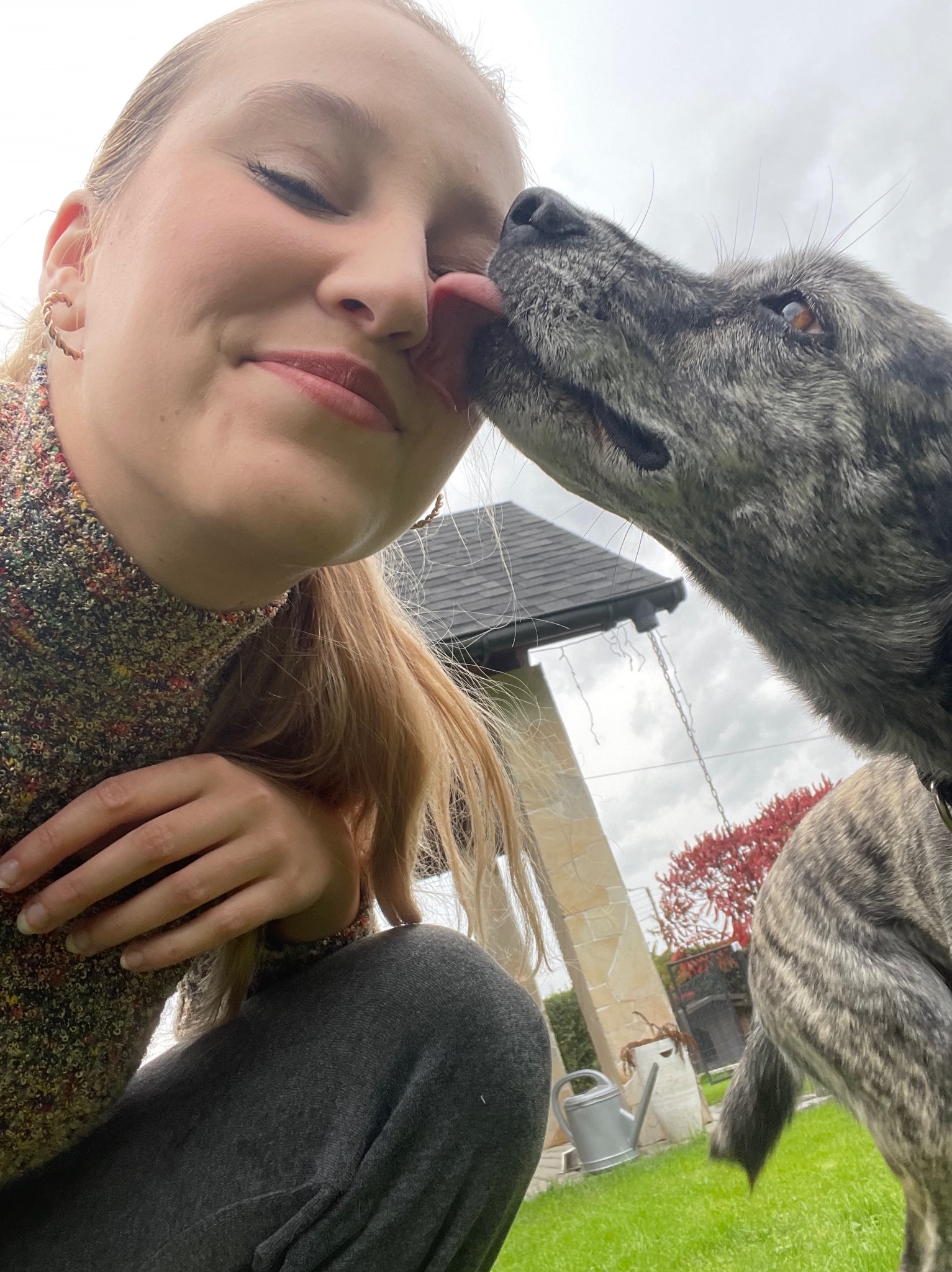 Kobieta z zamkniętymi oczami, której twarz liże duży, pręgowany pies na tle zielonego trawnika i zarysu budynku z szarym dachem.