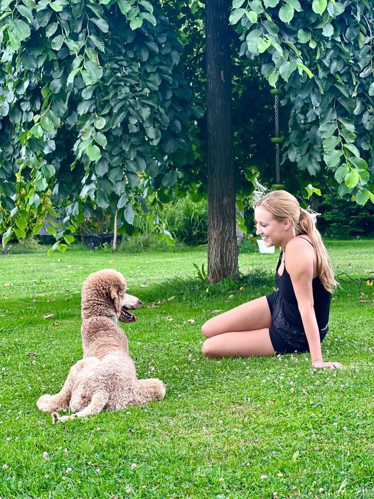 Kobieta siedzi na trawie obok pudla królewskiego w parku, oboje patrzą na siebie z uśmiechem.