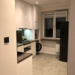 REMKAD - Nowoczesna, jasna kuchnia z białymi szafkami, drewnianym blatem, płytą gazową i czarną lodówką, widziana z perspektywy wejścia do pomieszczenia.