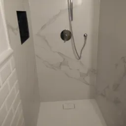 Nowoczesny prysznic walk-in z białymi płytkami imitującymi marmur i czarną wnęką na kosmetyki, widoczny odpływ liniowy.