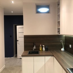 REMKAD - Nowoczesna kuchnia z białymi szafkami, drewnianym blatem i ciemnoniebieską ścianą w tle, oświetlona punktowymi lampami sufitowymi i lampką biurkową na blacie.