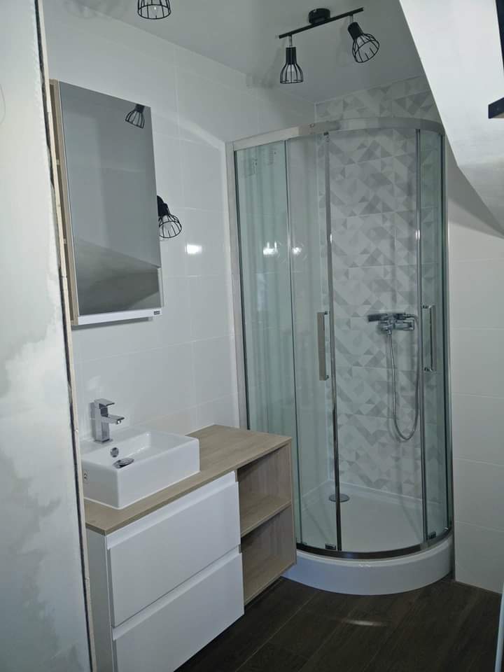 Nowoczesna łazienka z kabiną prysznicową narożną, umywalką nablatową na szafce z szufladami i półkami, oraz białymi płytkami na ścianach i ciemną podłogą z paneli.