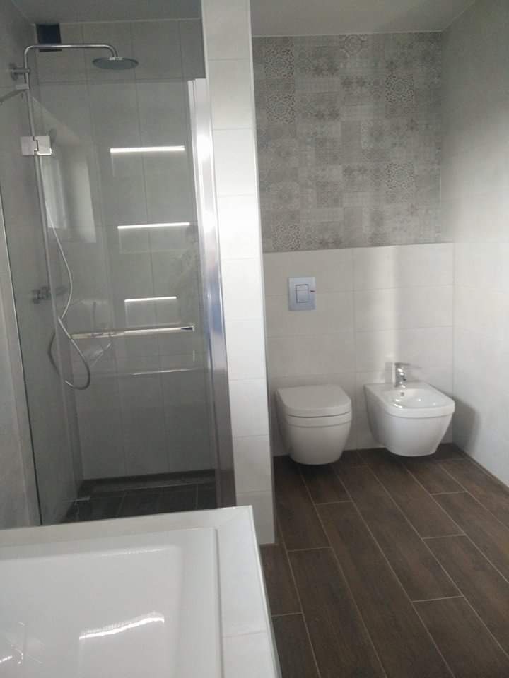 Nowoczesna łazienka z kabiną prysznicową typu walk-in, wiszącą toaletą i bidetem, wykończona płytkami imitującymi drewno na podłodze i szarymi płytkami dekoracyjnymi na ścianie powyżej białych płytek.