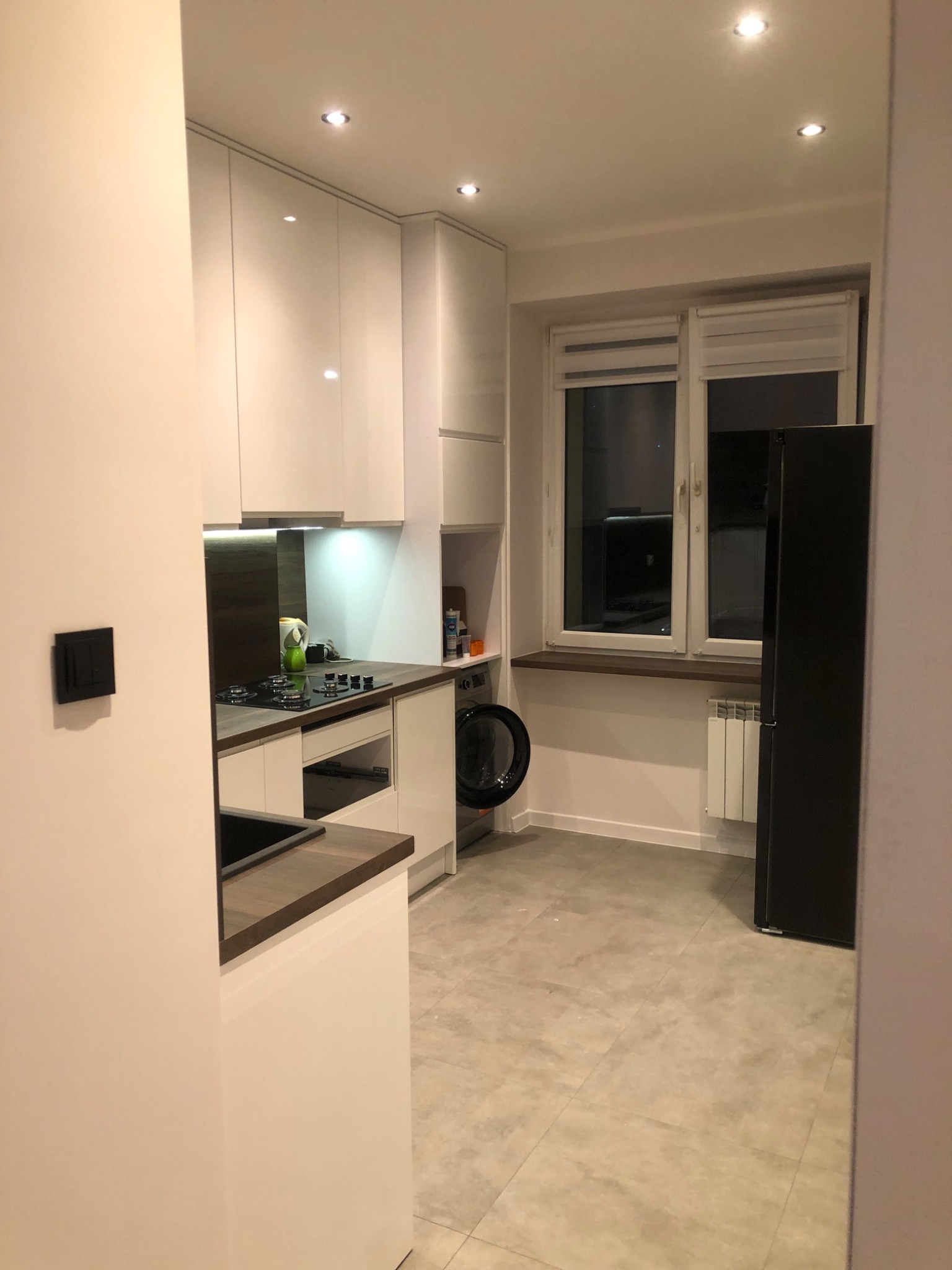 Nowoczesna, jasna kuchnia z białymi szafkami, drewnianym blatem, płytą gazową i czarną lodówką, widziana z perspektywy wejścia do pomieszczenia.