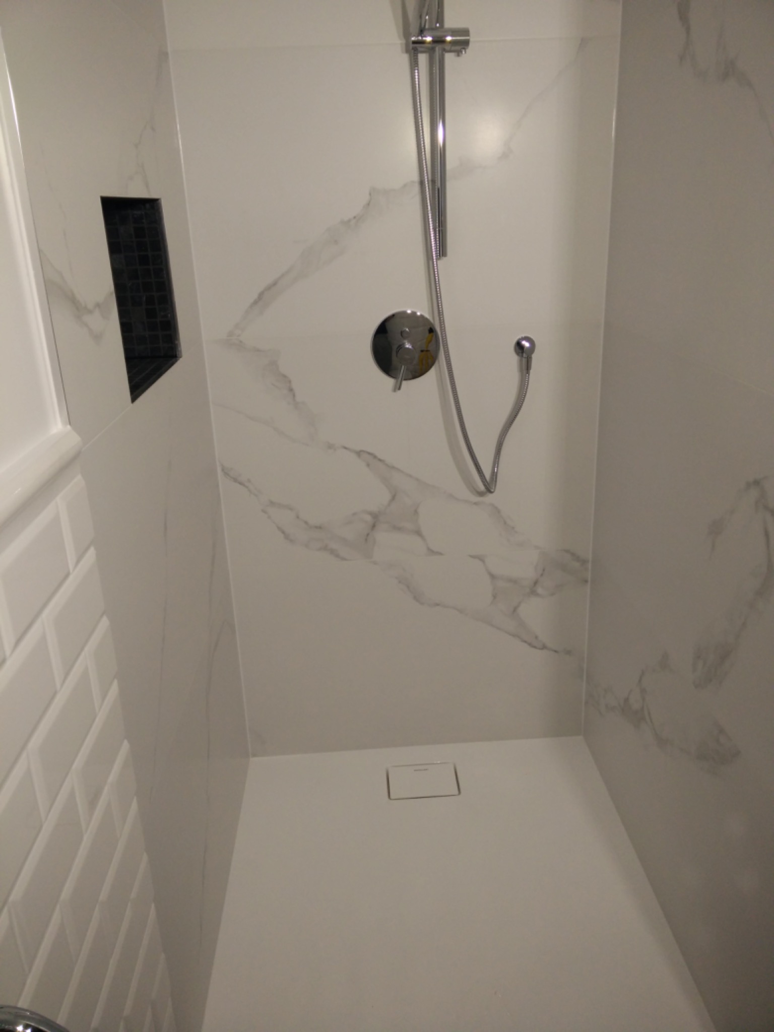 Nowoczesny prysznic walk-in z białymi płytkami imitującymi marmur i czarną wnęką na kosmetyki, widoczny odpływ liniowy.