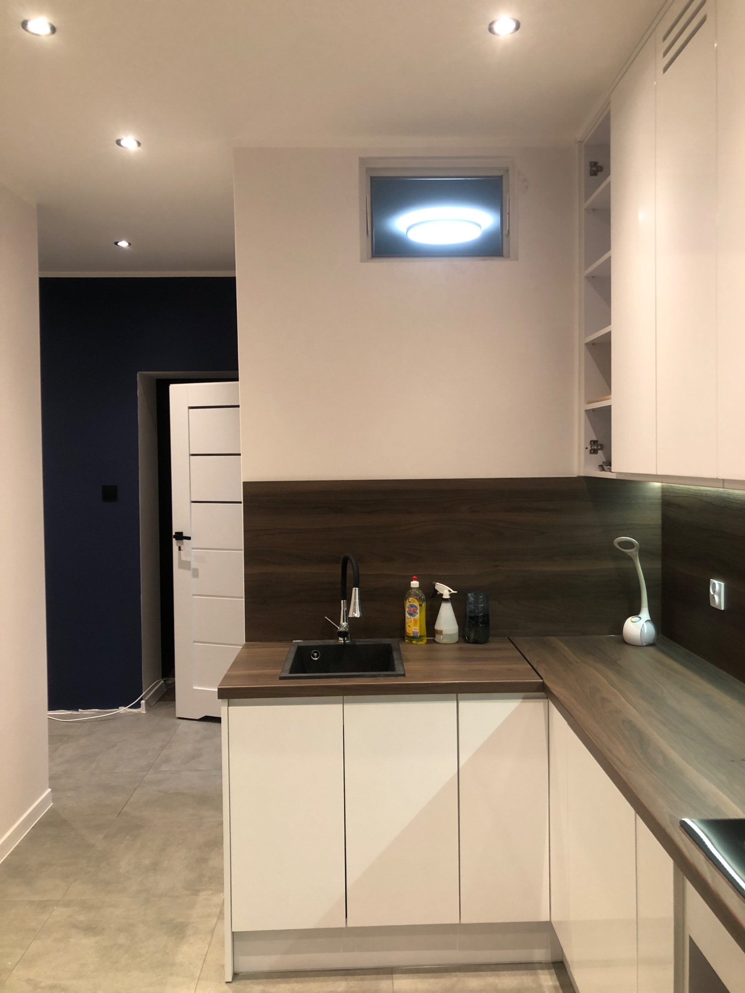 Nowoczesna kuchnia z białymi szafkami, drewnianym blatem i ciemnoniebieską ścianą w tle, oświetlona punktowymi lampami sufitowymi i lampką biurkową na blacie.