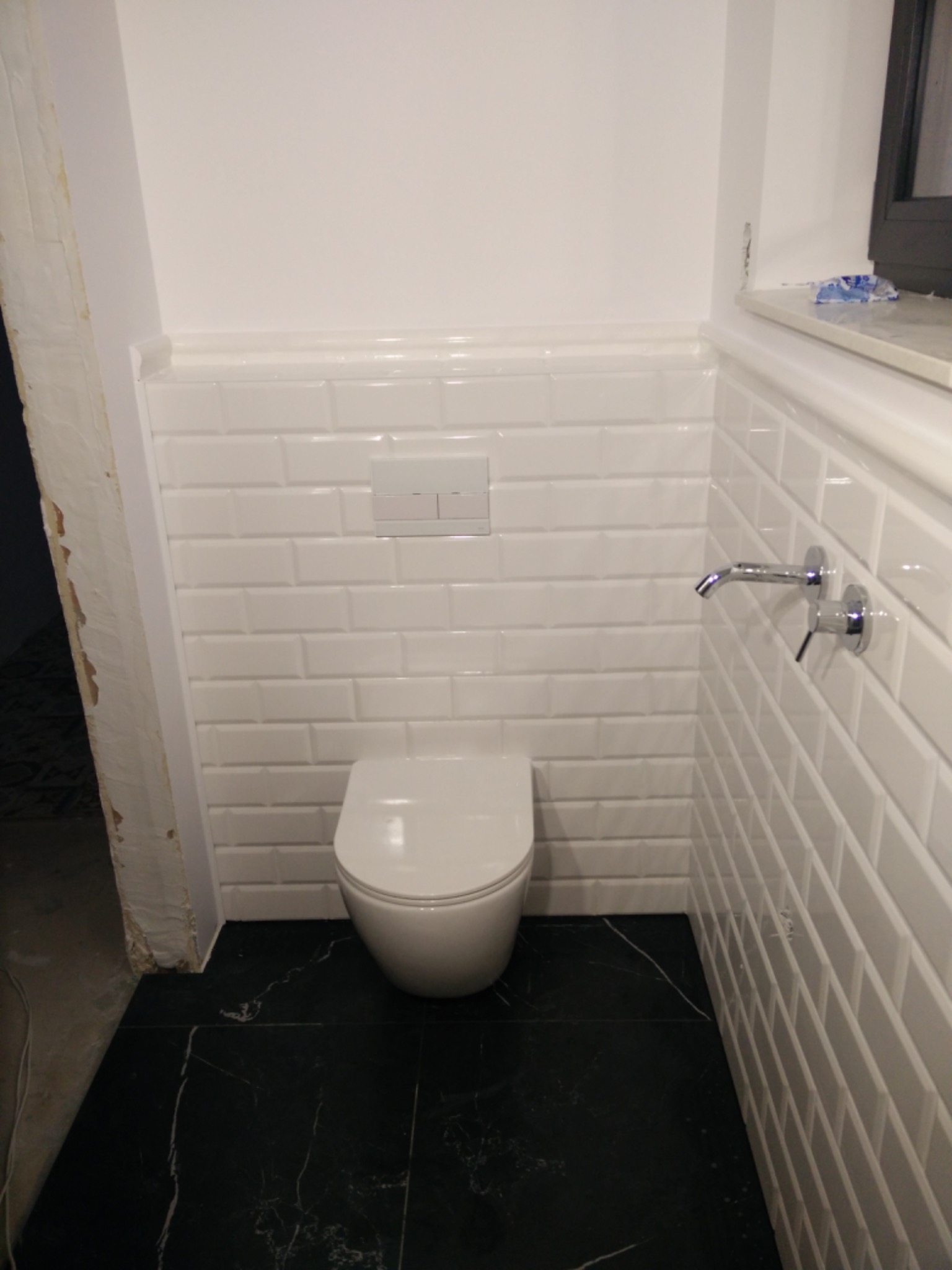 Nowoczesna toaleta z białymi, prostokątnymi płytkami ułożonymi w cegiełkę i czarną marmurową podłogą, widoczne chromowane baterie ścienne i fragment nie wykończonego otworu drzwiowego.