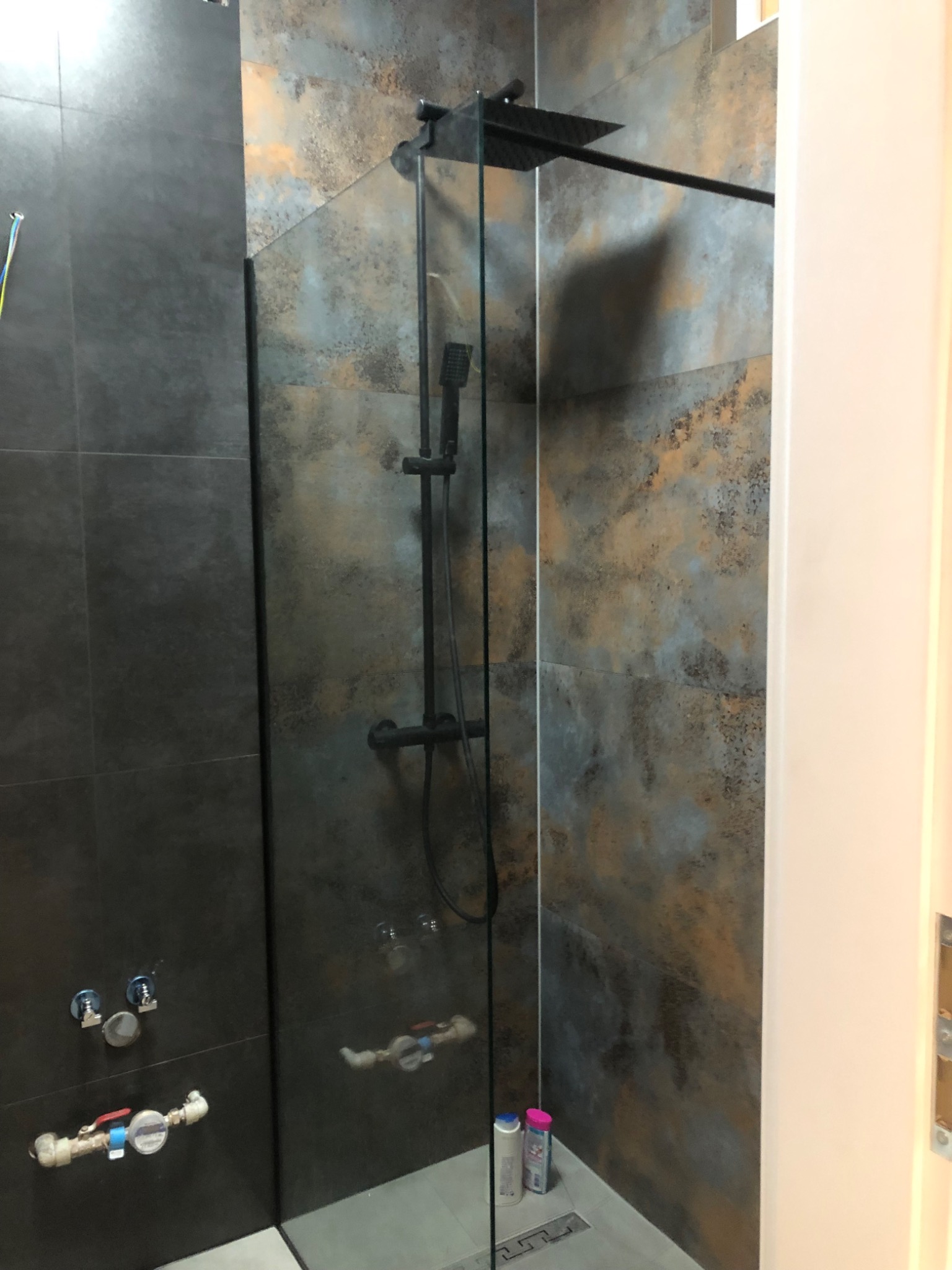 Nowoczesna kabina prysznicowa z czarną armaturą i szklaną ścianką, wykończona płytkami imitującymi rdzawy metal, widoczne przyłącza wodne na ścianie.