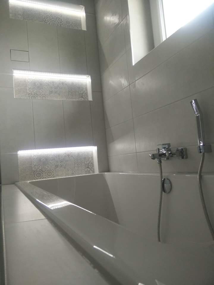 Minimalistyczna łazienka z białą wanną, szare płytki z dekoracyjnymi wnękami podświetlonymi LED, chromowana bateria i słuchawka prysznicowa.