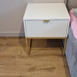 Biała szafka nocna z ryflowanym frontem szuflady i złotymi n&oacute;żkami, stojąca na drewnianej podłodze obok szarego ł&oacute;żka z r&oacute;żową pościelą.