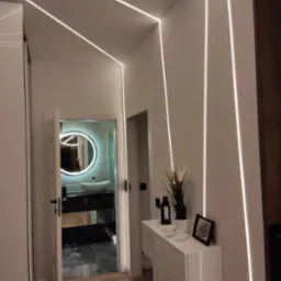 Nowoczesny korytarz z oświetleniem LED wzdłuż ścian i sufitu, widok na łazienkę z okrągłym lustrem podświetlanym LED, biała szafa przesuwna po lewej, drewnopodobna podłoga, biała konsola...