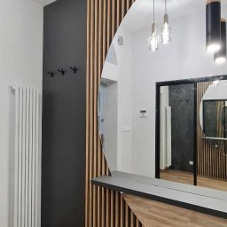 MEBLE NIKOMAX - Nowoczesny przedpokój z drewnianymi lamelami na ścianie, owalnym lustrem i czarną ścianą z wieszakami, oświetlenie wiszące.