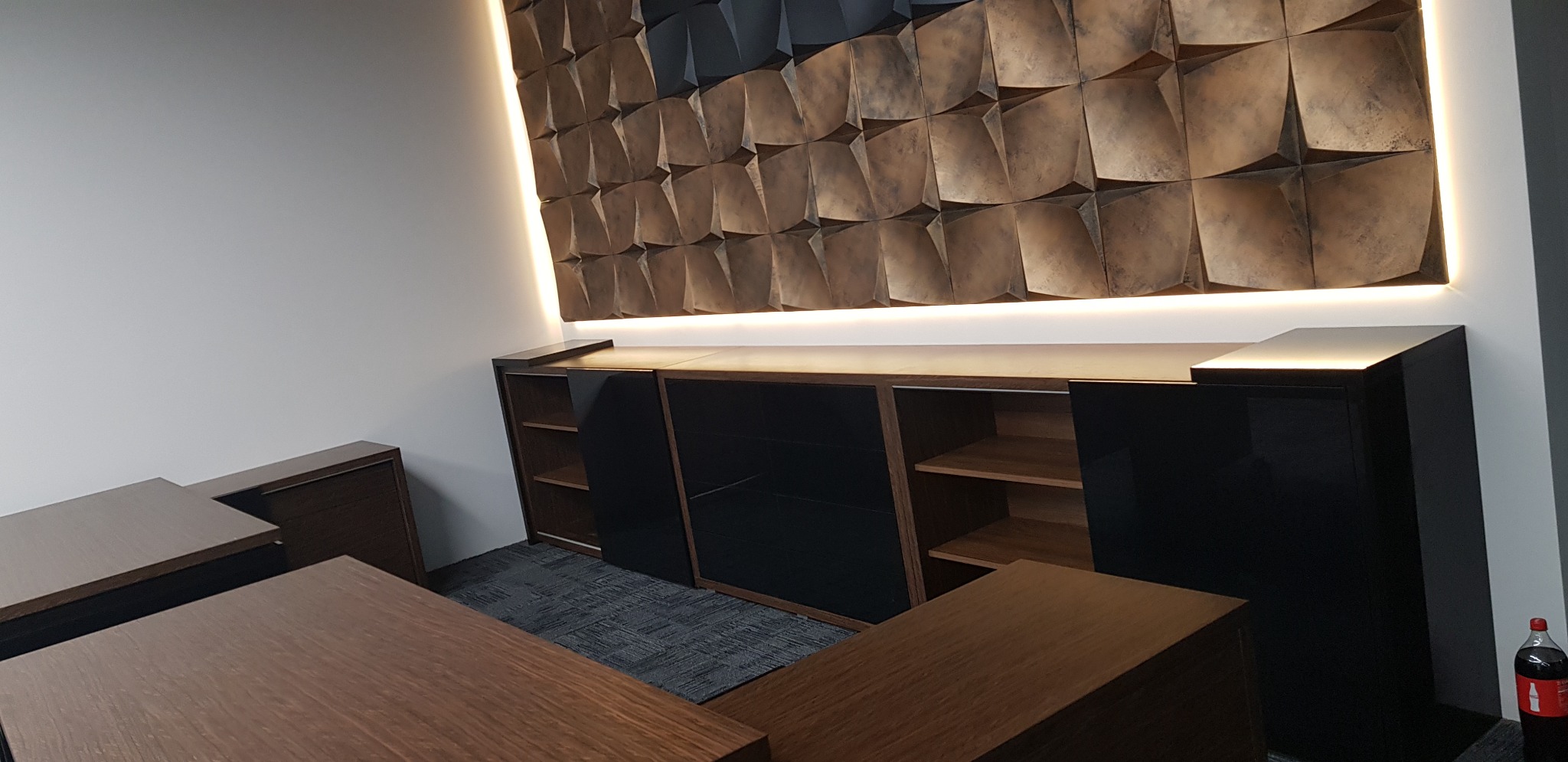 Nowoczesna zabudowa meblowa z czarnymi frontami i drewnianymi elementami, podświetlona listwą LED, na tle dekoracyjnej ściany z geometrycznym wzorem.