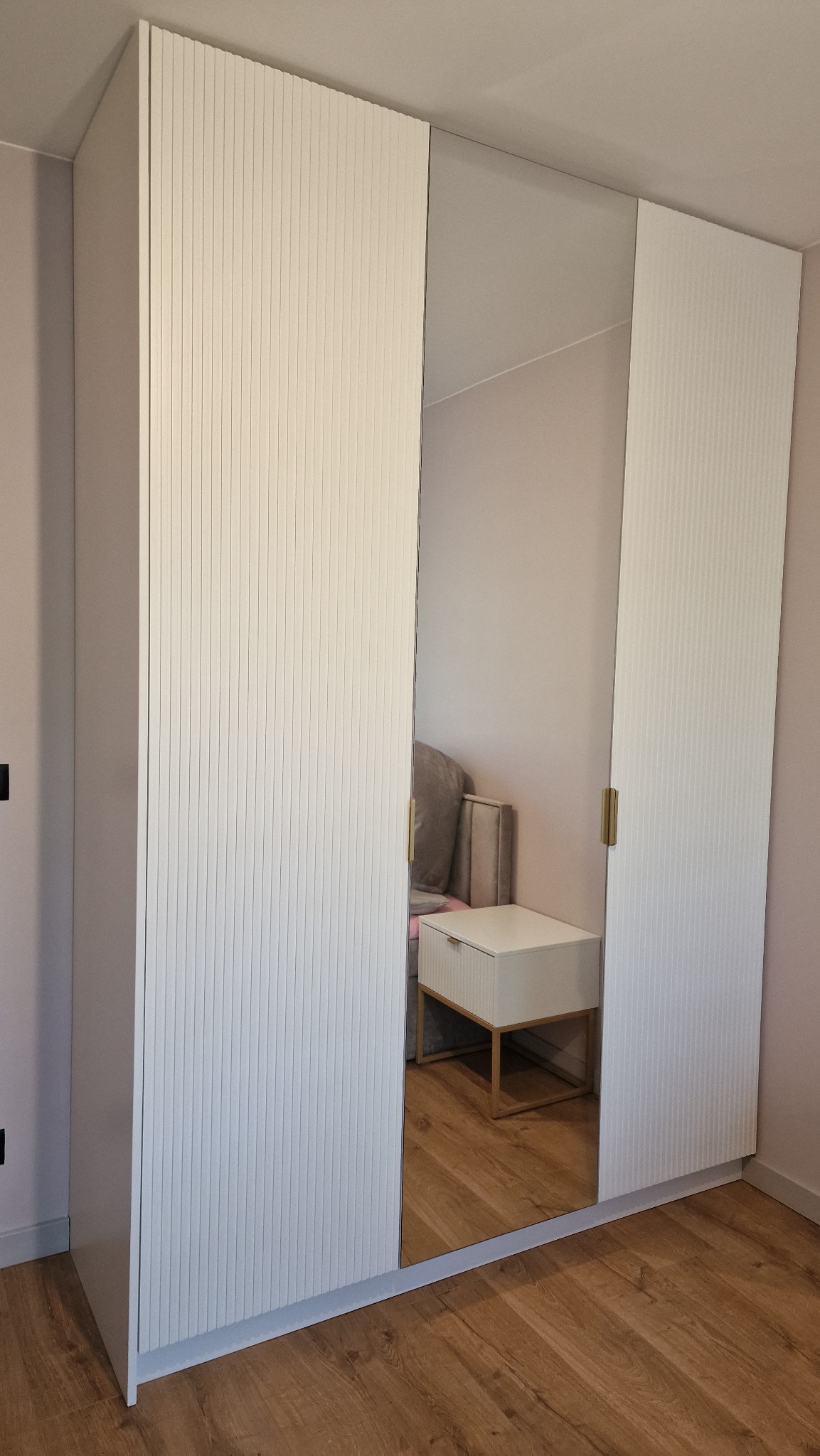 Biała szafa z lustrzanym frontem, ryflowane drzwi, złote uchwyty, odbicie szafki nocnej i wezgłowia łóżka.