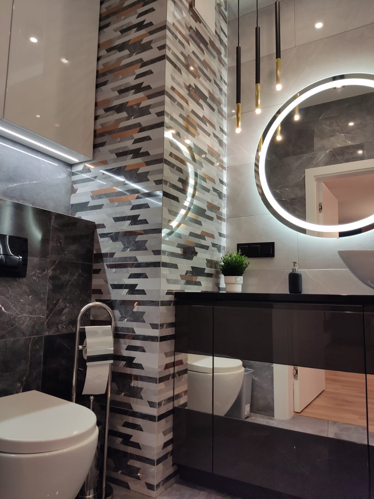 Nowoczesna łazienka z podświetlanym lustrem, kolumną z mozaiki i czarną szafką pod umywalką, widoczne wiszące lampy i toaleta.