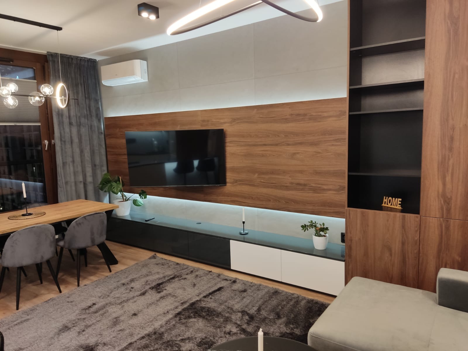 Nowoczesny salon z zabudową meblową z forniru drewnopodobnego, podświetleniem LED, szarą sofą i dywanem, oraz stołem jadalnym z szarymi krzesłami.