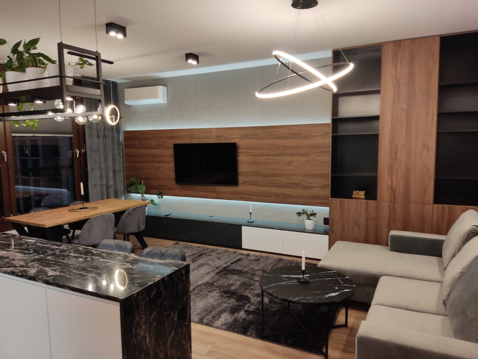 Nowoczesne wnętrze salonu z elementami drewna: ściana z telewizorem, zabudowa meblowa, stół jadalny, sofa narożna i wyspa kuchenna z blatem z czarnego marmuru.